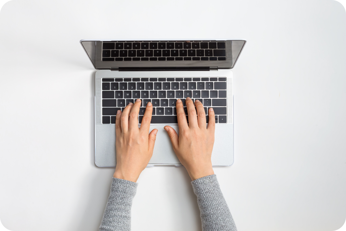 Widok z góry na dłonie piszące na klawiaturze srebrnego laptopa ustawionego na jasnym tle.