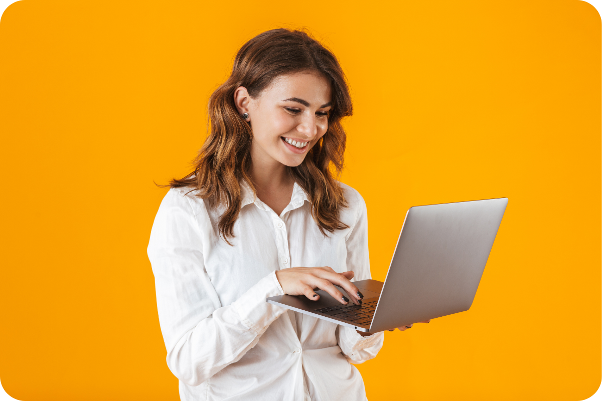 Uśmiechnięta kobieta w białej koszuli pracuje na laptopie na intensywnie pomarańczowym tle.